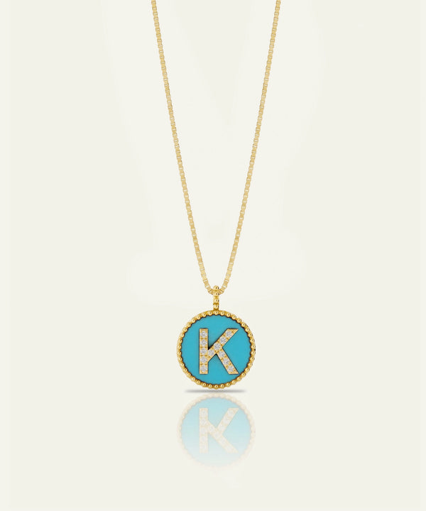 A gold chain necklace with a round pendant featuring a turquoise blue background and a raised gold letter 'K' outlined with small clear stones, shown on a light cream background with a soft reflection below.