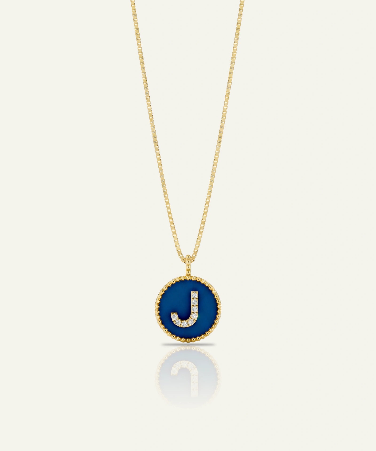 A gold chain necklace with a circular pendant featuring a deep blue enamel background and a raised gold letter 'J' outlined with small clear stones, shown on a light cream background with a soft reflection below.
