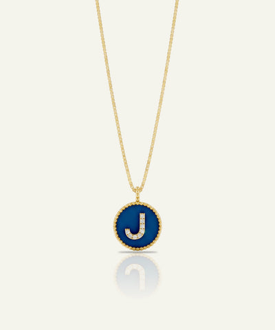 A gold chain necklace with a circular pendant featuring a deep blue enamel background and a raised gold letter 'J' outlined with small clear stones, shown on a light cream background with a soft reflection below.