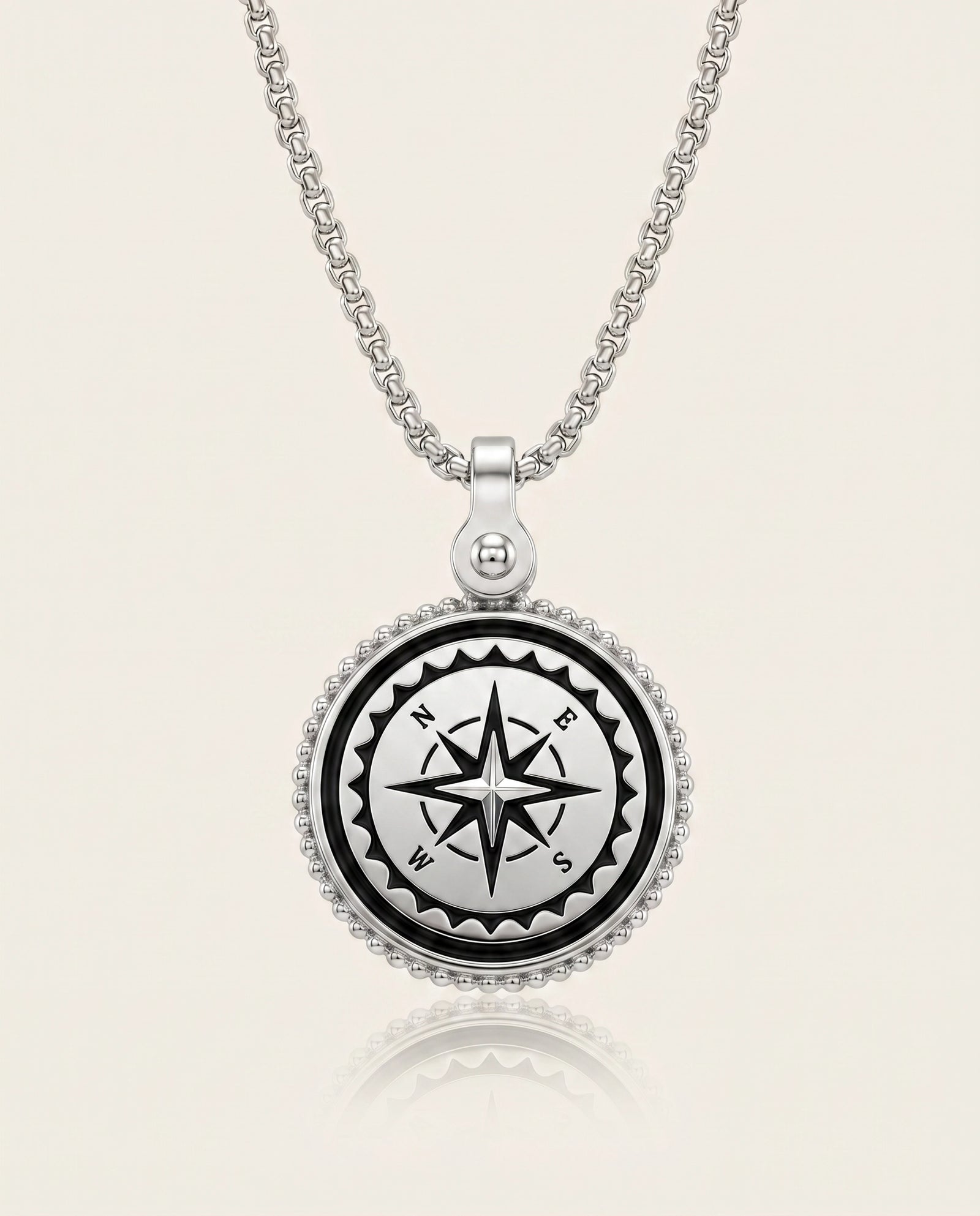 A silver compass pendant necklace on a matching chain, featuring a black compass rose design in the center with white lettering, surrounded by a beaded silver border, set against a light cream background.