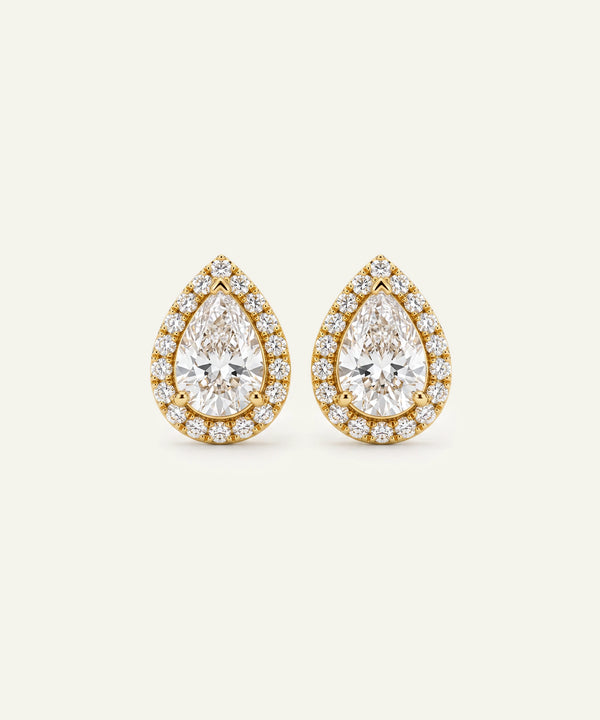 A pair of pear-shaped diamond stud earrings in yellow gold, featuring a large central pear-cut diamond surrounded by a halo of smaller round diamonds, set against a light cream background.