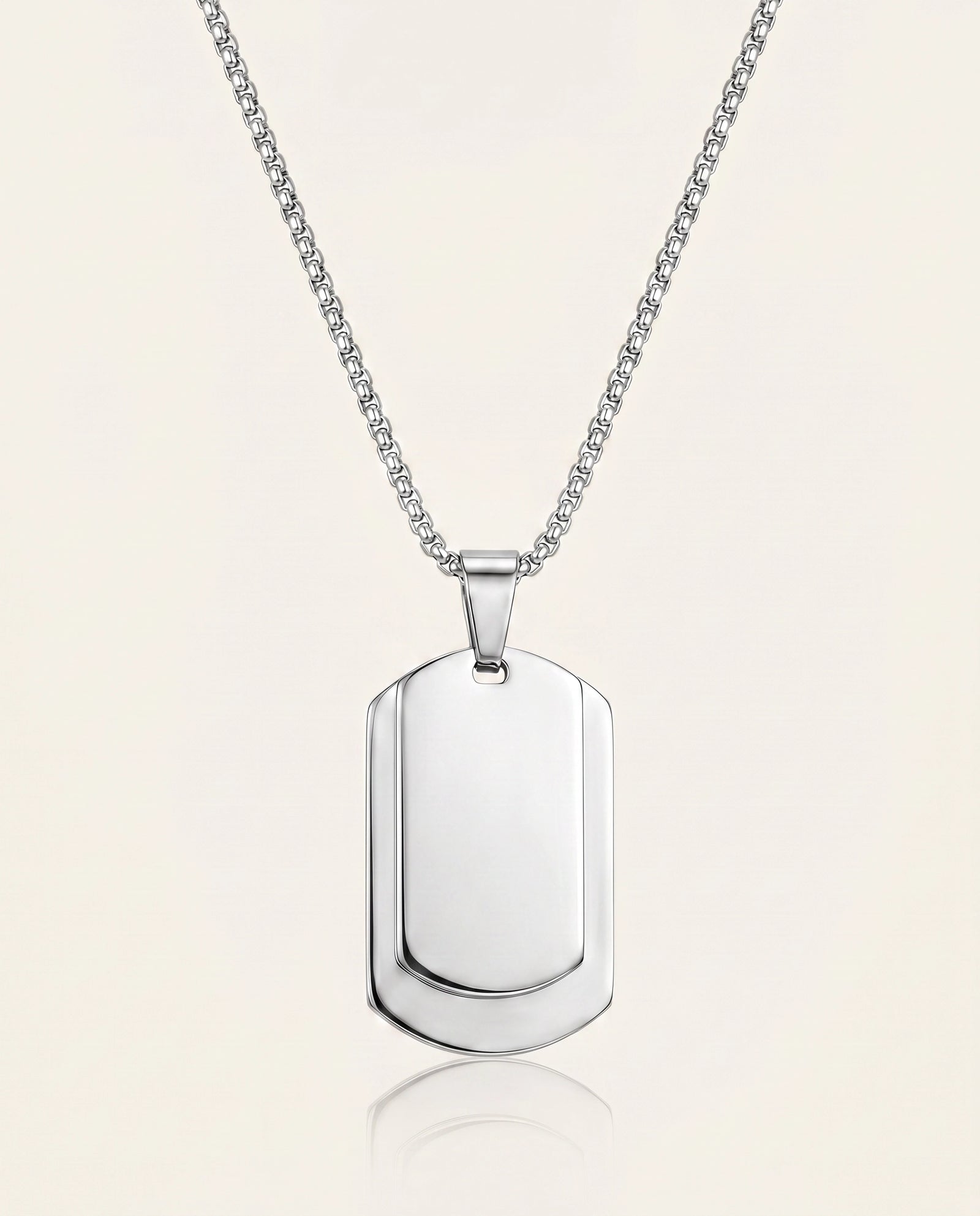 A silver dog tag pendant necklace on a delicate silver chain, presented on a light cream background with a soft reflection below.