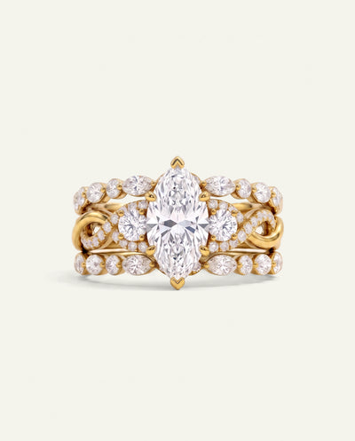 A yellow gold ring set featuring a prominent marquise-cut center stone surrounded by round and oval diamonds, with additional diamond accents along the band, photographed on a light cream background.