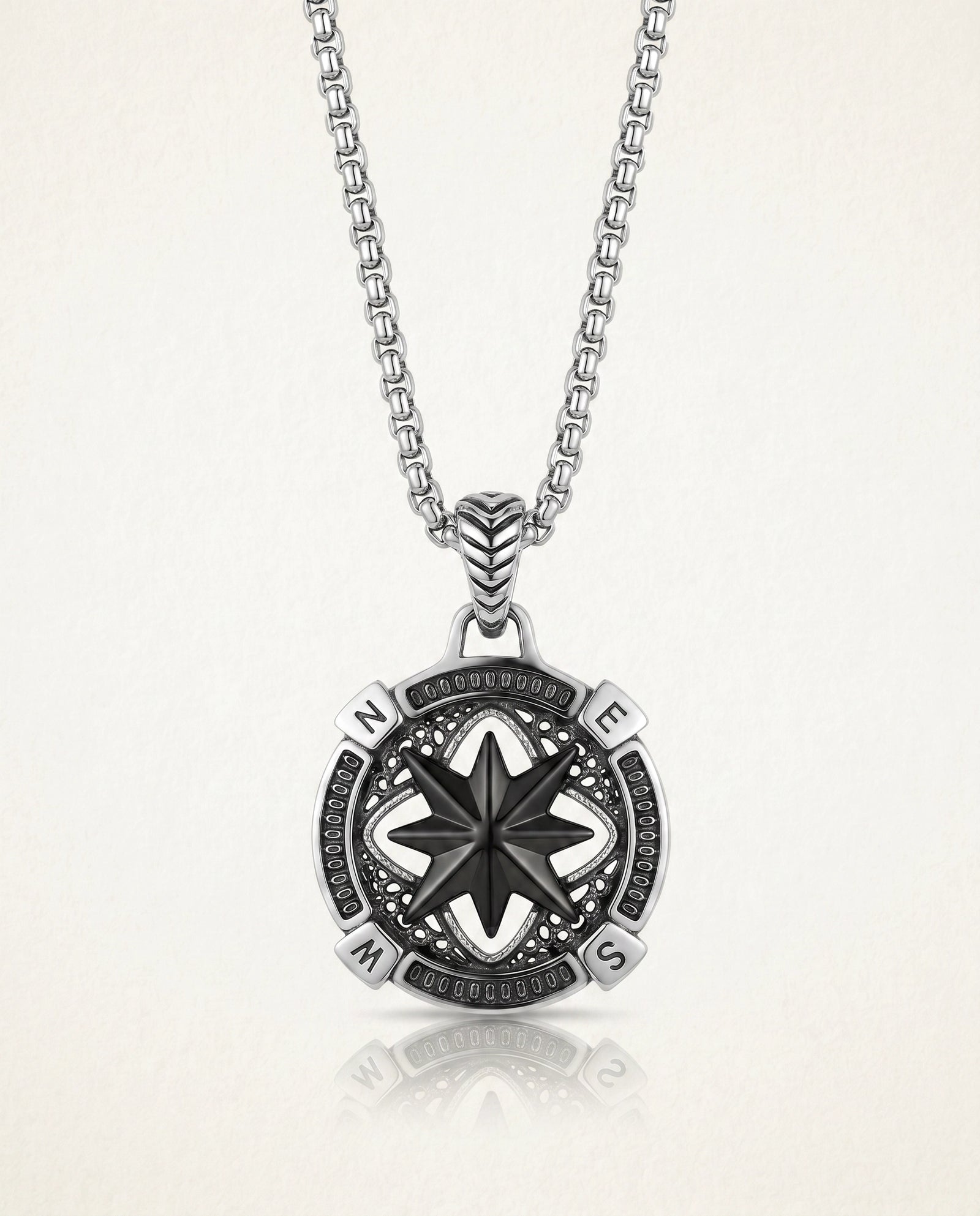 A silver compass pendant necklace on a matching chain, displayed against a light beige background with a soft reflection below.