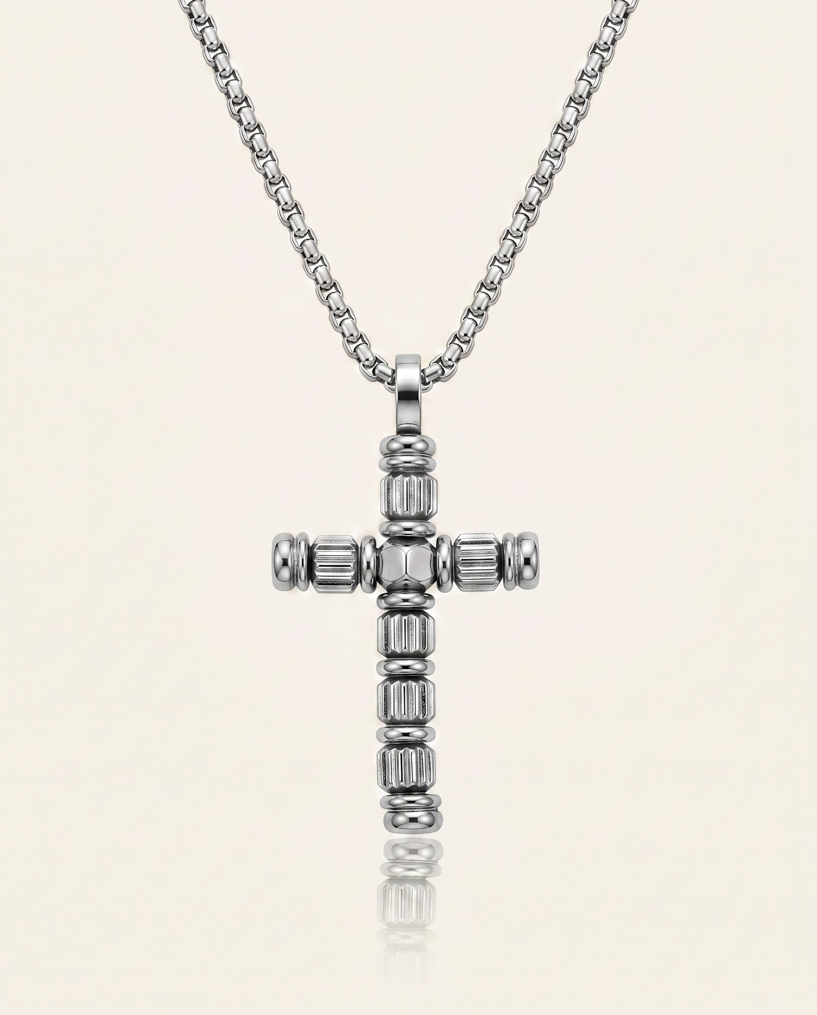 A silver cross pendant necklace with a beaded, textured design, suspended from a matching silver box chain against a light cream background.