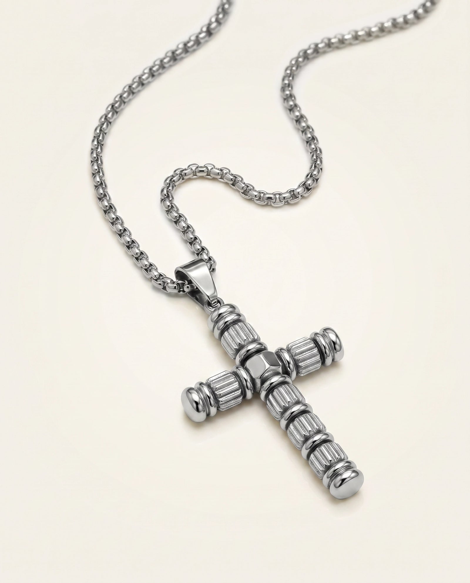 A silver-toned cross pendant necklace with a textured, ribbed design and a matching box chain, displayed on a light cream background.