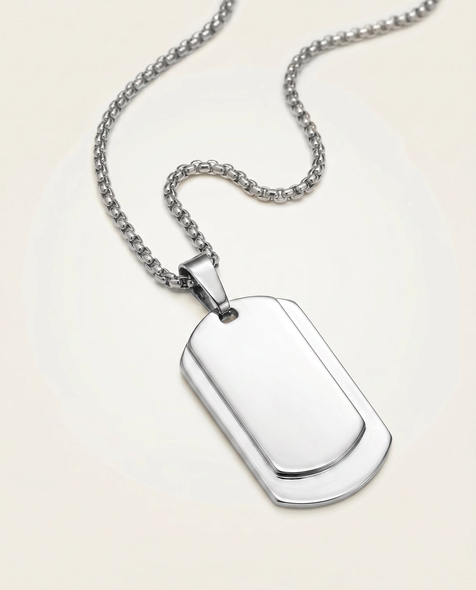 A polished silver-toned metal dog tag pendant featuring a layered, contoured center plate, hanging from a matching silver box chain against a solid light cream background.