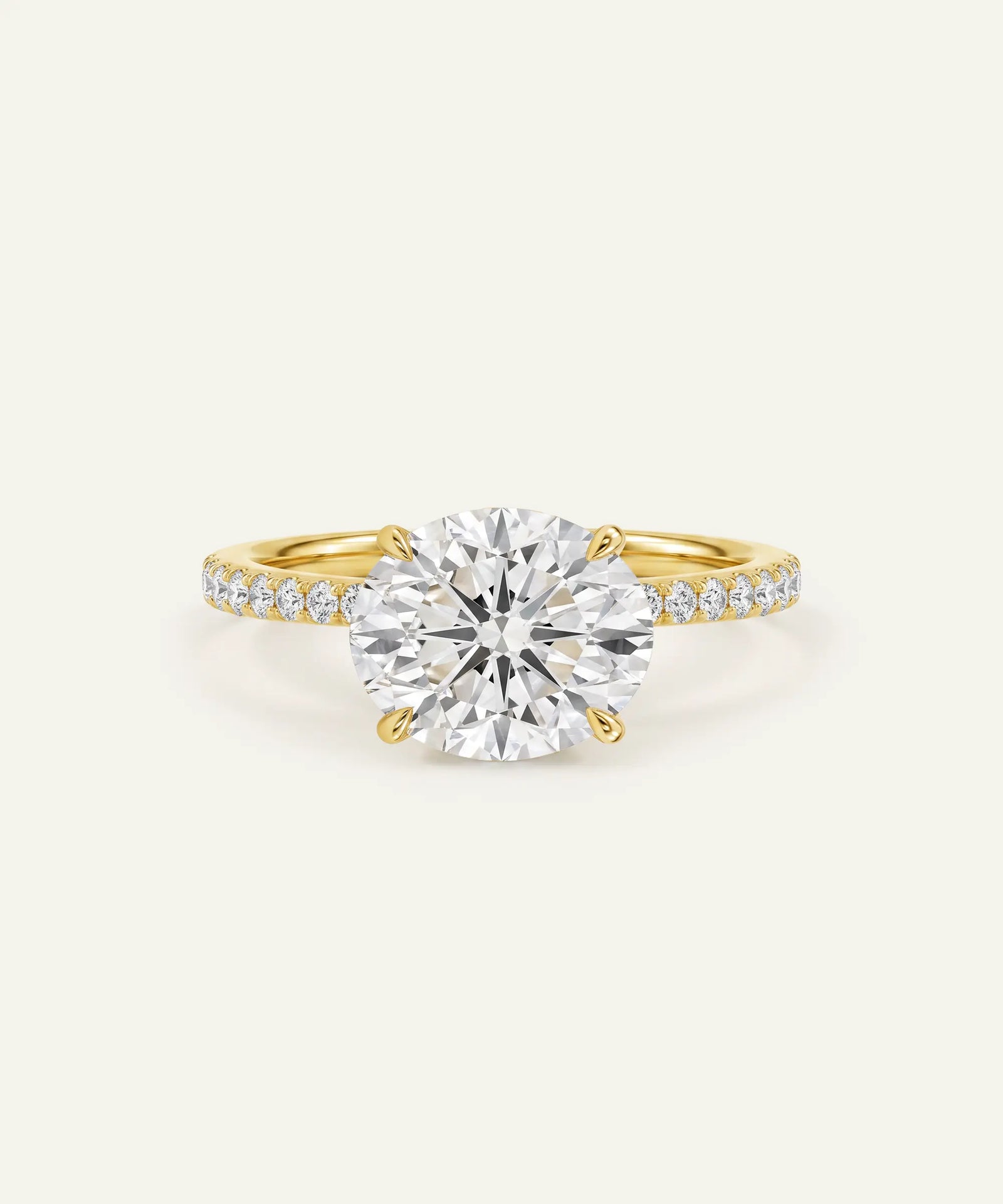 A yellow gold solitaire engagement ring featuring a large round brilliant-cut center stone and a pavé-set diamond band, shown on a light cream background.