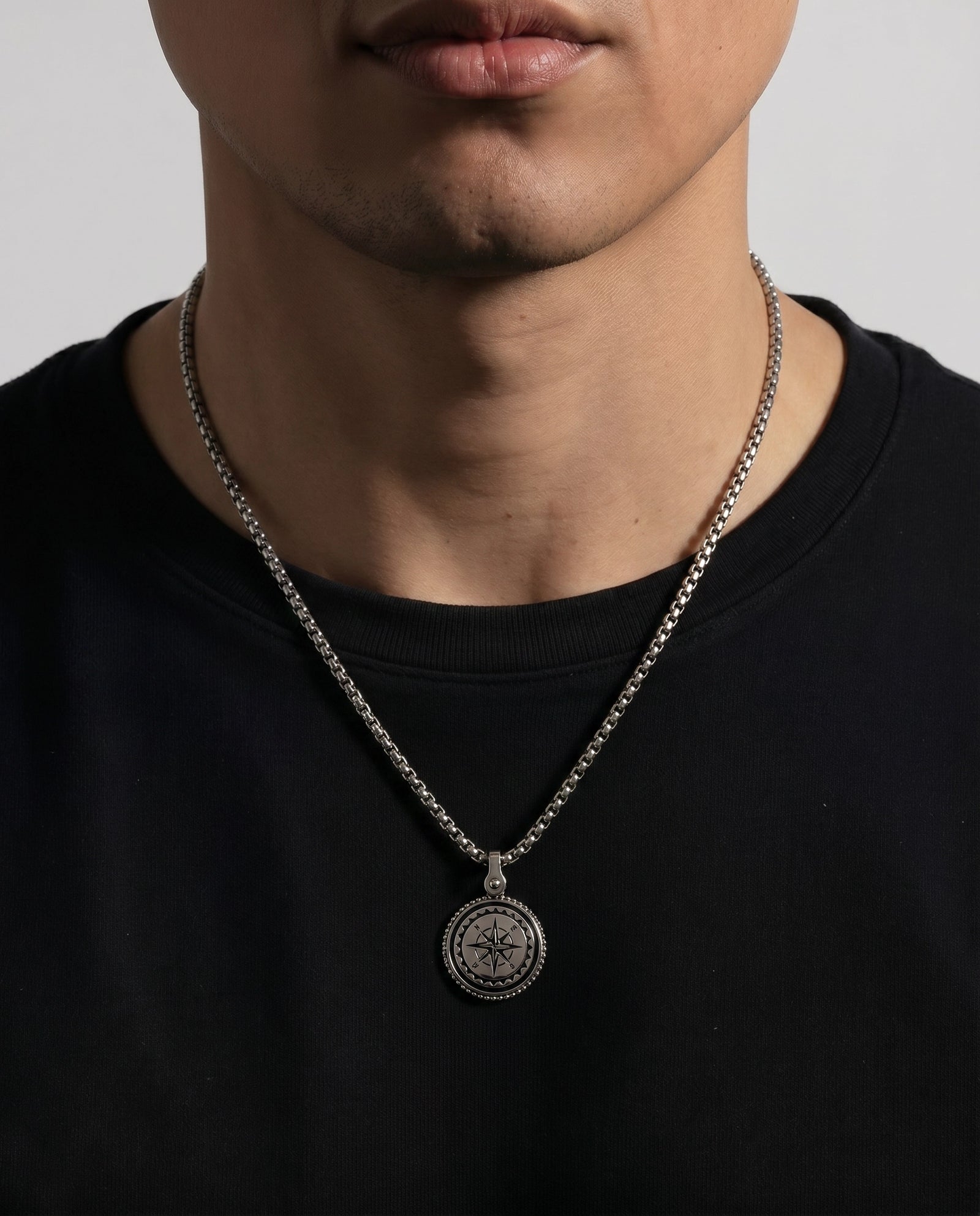 A close-up of a man wearing a silver chain necklace with a circular compass pendant, paired with a black crewneck t-shirt against a light gray background.