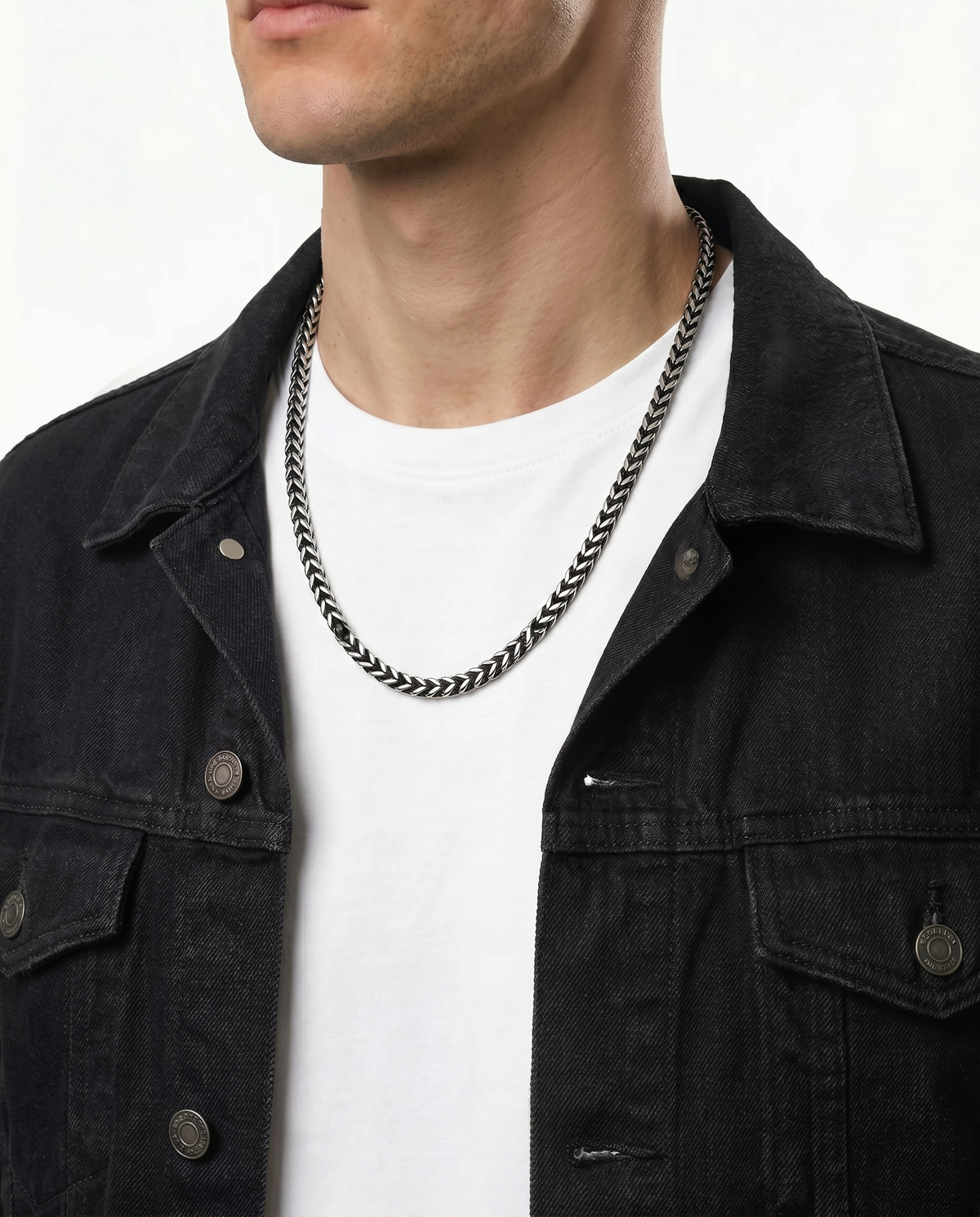 A close-up of a man wearing a silver-toned chain necklace over a white t-shirt and a black denim jacket.