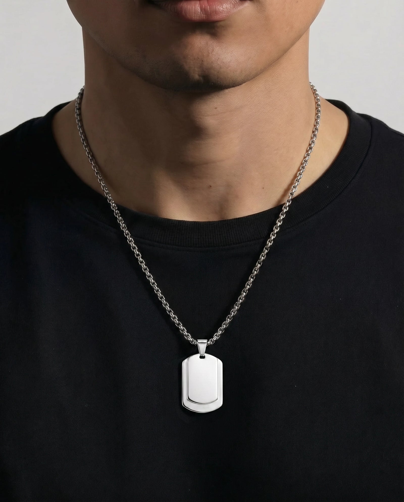 A close-up of a man wearing a silver dog tag pendant necklace on a black t-shirt against a light gray background.