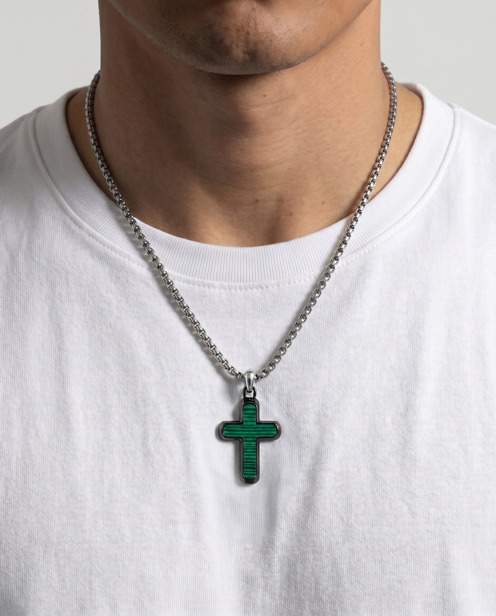 A close-up of a person wearing a silver chain necklace with a textured green cross pendant, paired with a white crew-neck t-shirt.