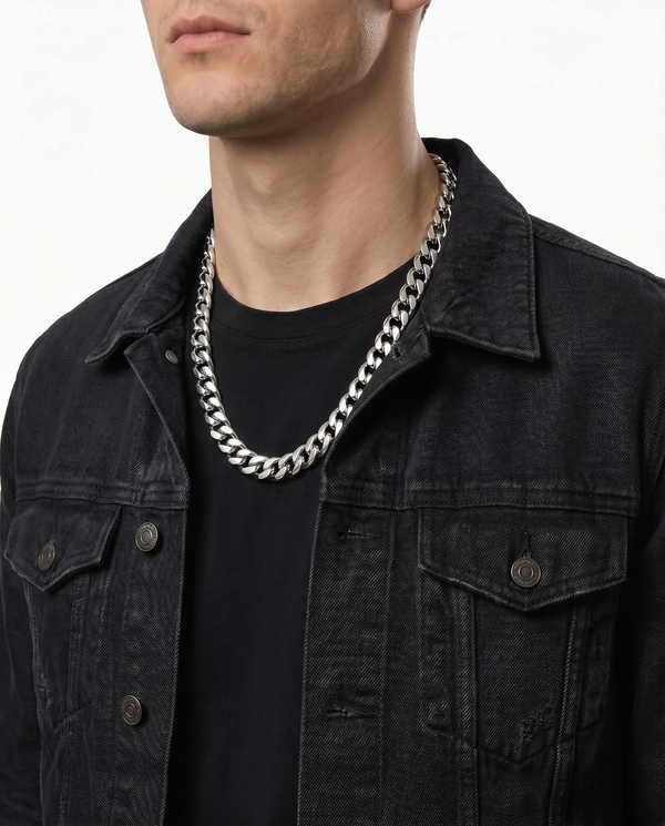 A close-up of a man wearing a chunky silver chain necklace over a black t-shirt and a dark denim jacket, against a plain white background.
