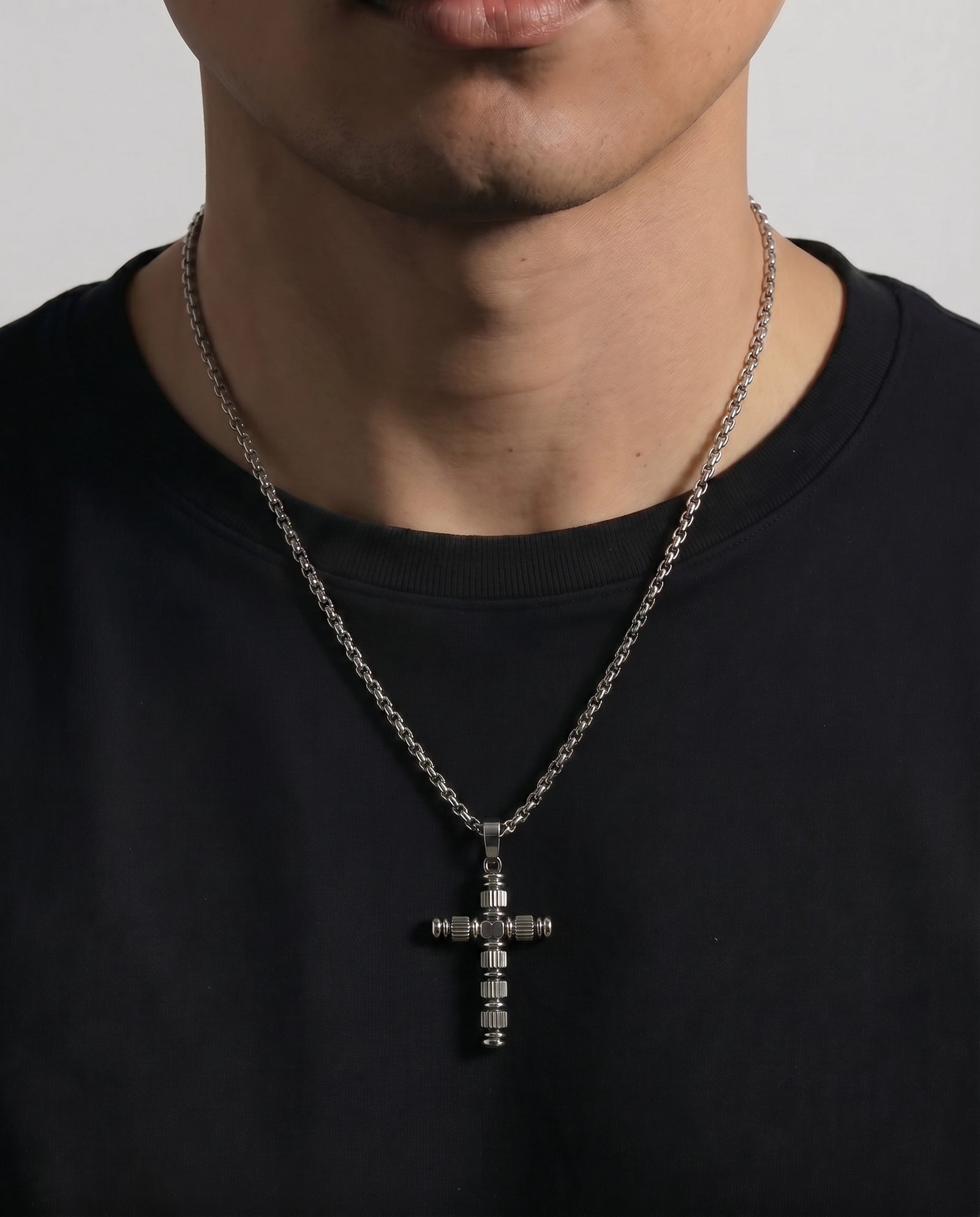 A close-up of a person wearing a silver-toned cross pendant necklace with clear stones, paired with a black crew-neck t-shirt against a light gray background.