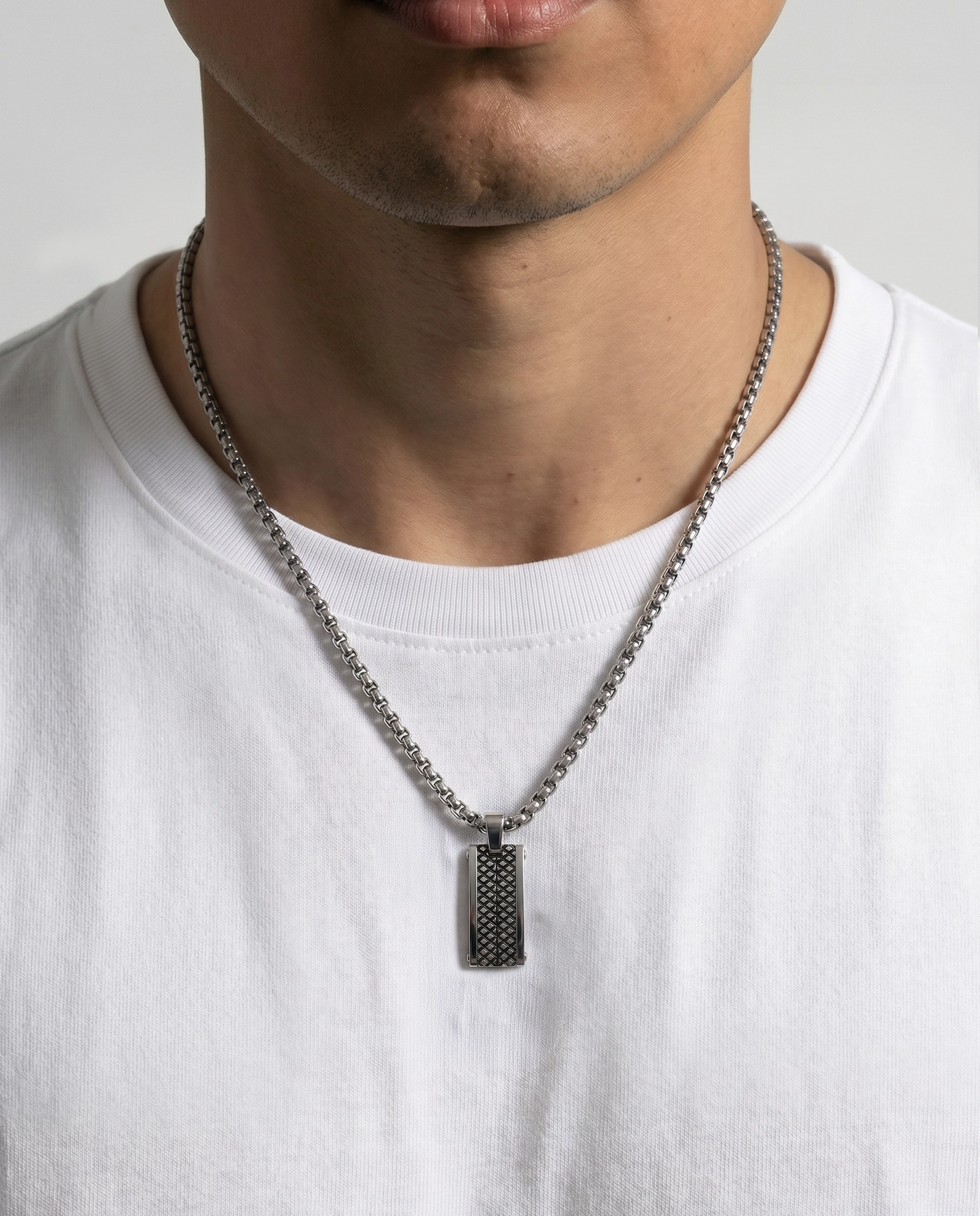 A close-up of a man wearing a silver chain necklace with a textured rectangular pendant, paired with a white crew-neck t-shirt.