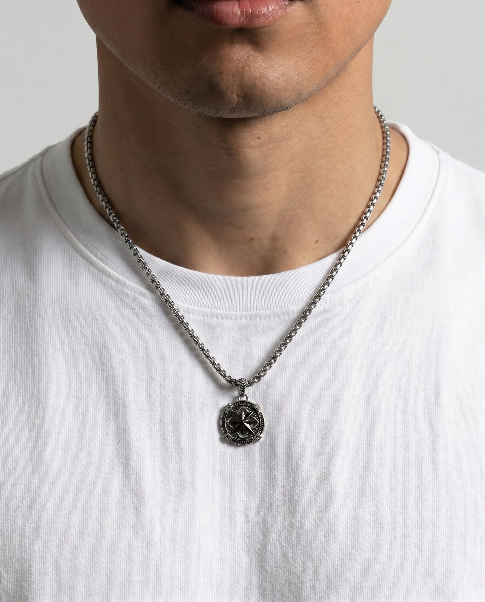 A close-up of a man wearing a dark metal pendant necklace with a circular emblem, styled over a white t-shirt
