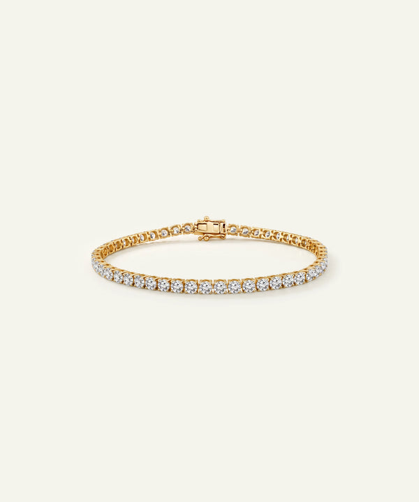 A gold tennis bracelet featuring a single row of clear round-cut gemstones on one side and a polished gold chain on the other, displayed on a light cream background.