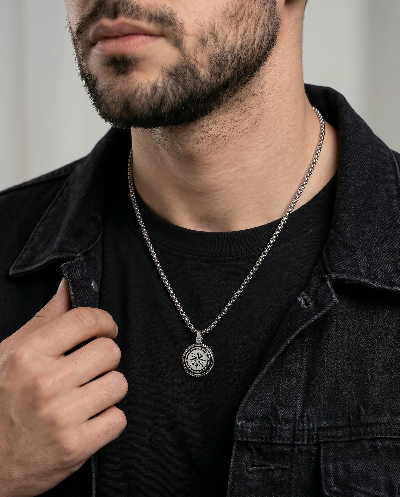 A close-up of a man wearing a silver compass pendant necklace over a black t-shirt and dark denim jacket.