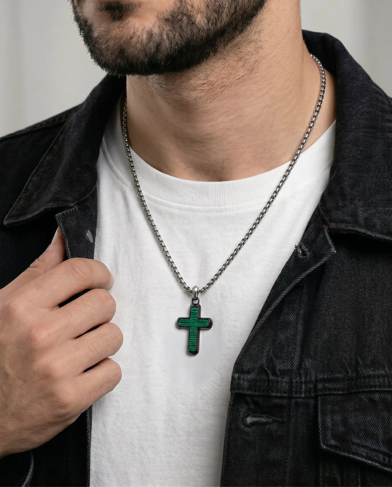 A close-up of a man wearing a silver chain necklace with a green cross pendant, styled over a white t-shirt and a dark denim jacket.