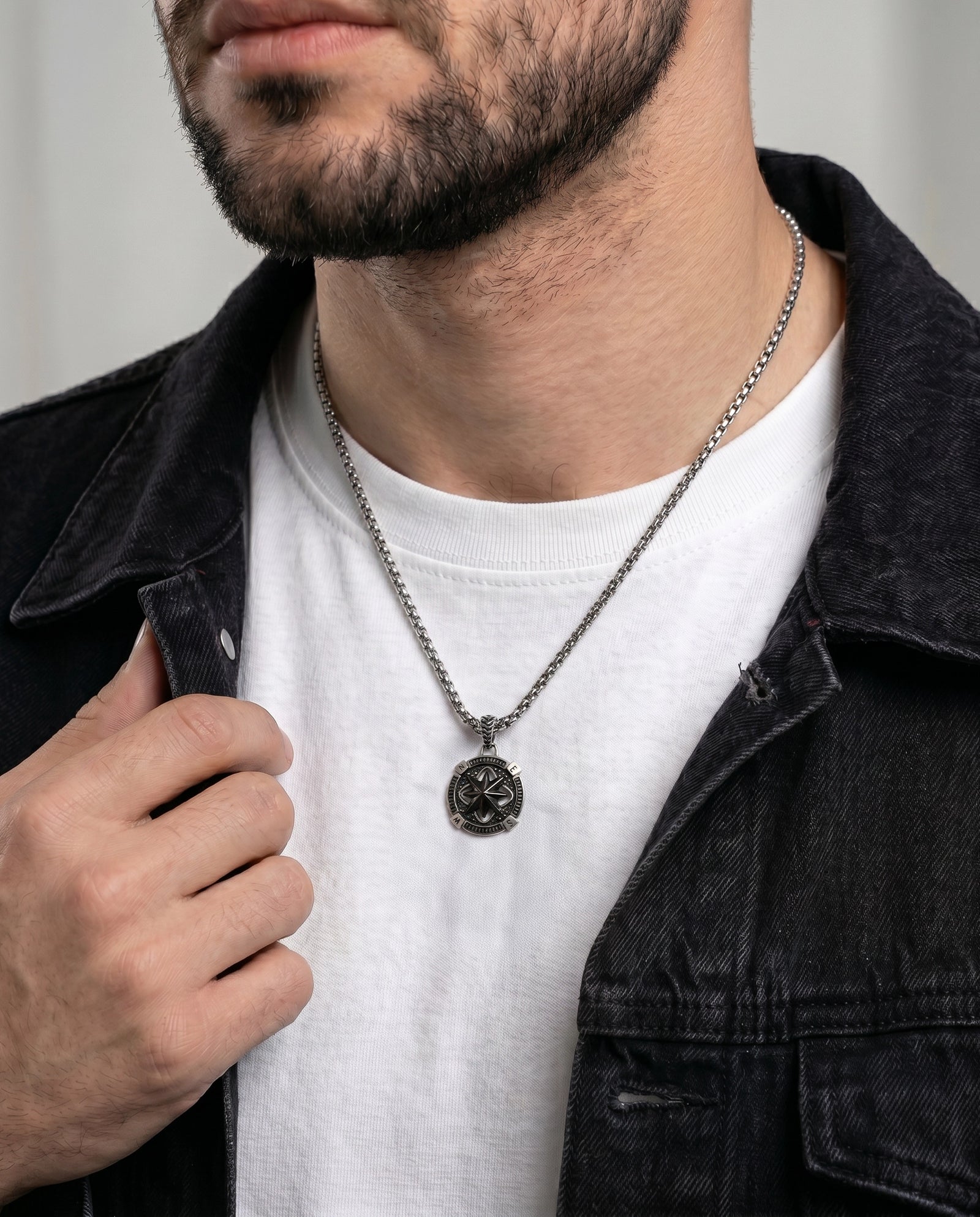 A close-up of a man wearing a dark metal pendant necklace with a circular emblem, styled over a white t-shirt and a dark denim jacket.