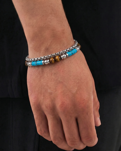 A close-up of a person's wrist and hand wearing two beaded bracelets. One bracelet is made of silver-toned metal beads, and the other features bright turquoise blue beads with silver spacer beads and a single brown tiger's eye bead. - alternate view