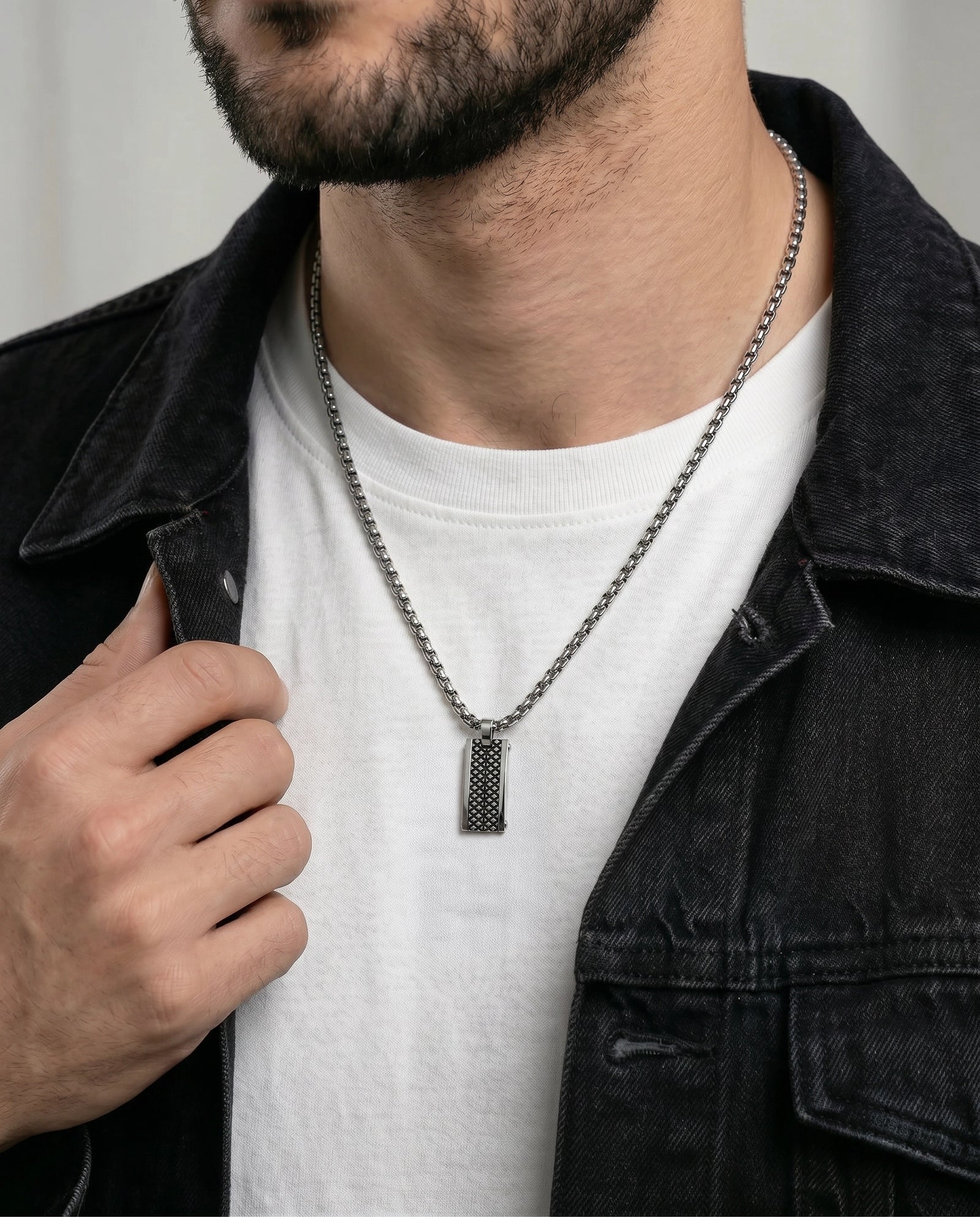 A close-up of a man wearing a dark metal chain necklace with a textured rectangular pendant, styled over a white t-shirt and a dark denim jacket.