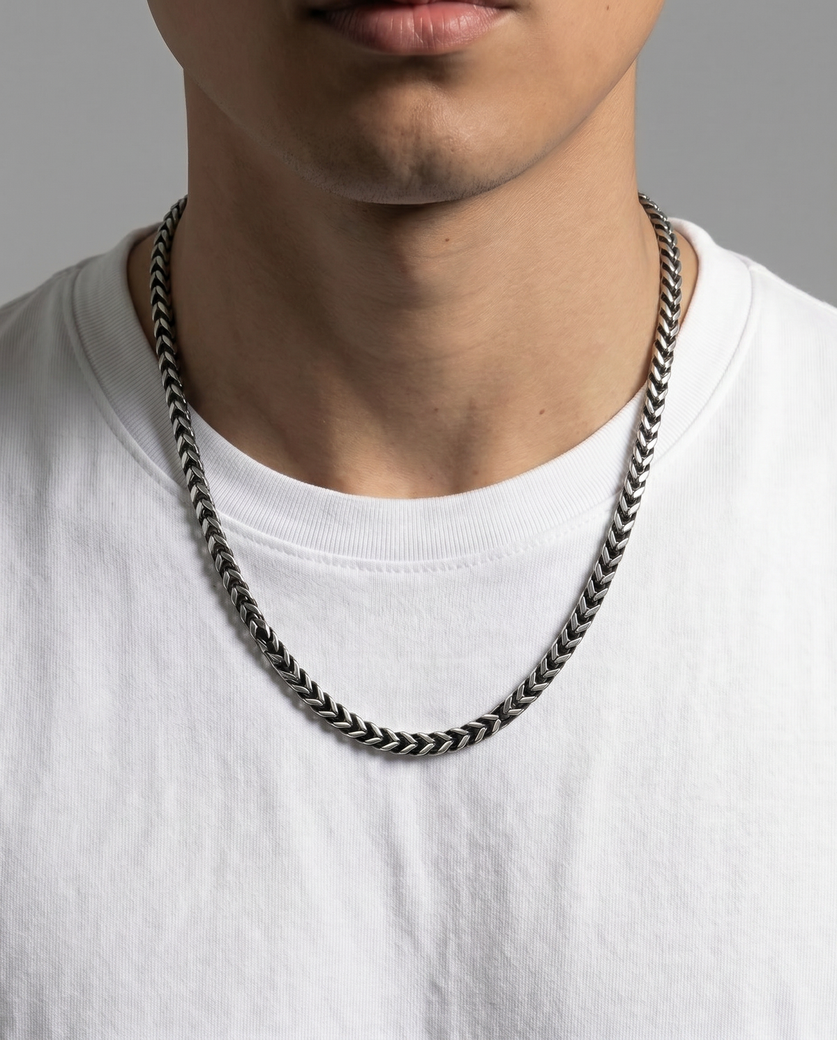 A close-up of a man wearing a thick, braided silver chain necklace over a white t-shirt, against a neutral gray background.