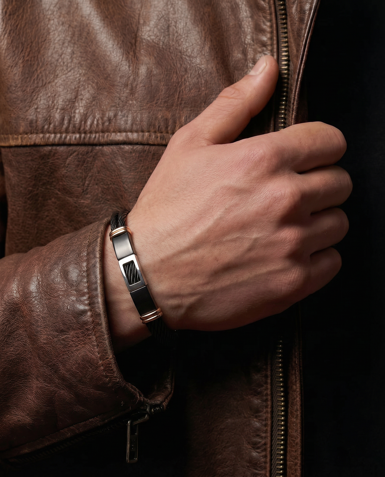 A close-up of a man's hand wearing a sleek black and silver metal bracelet with a geometric pattern, resting on his brown leather jacket zipper.