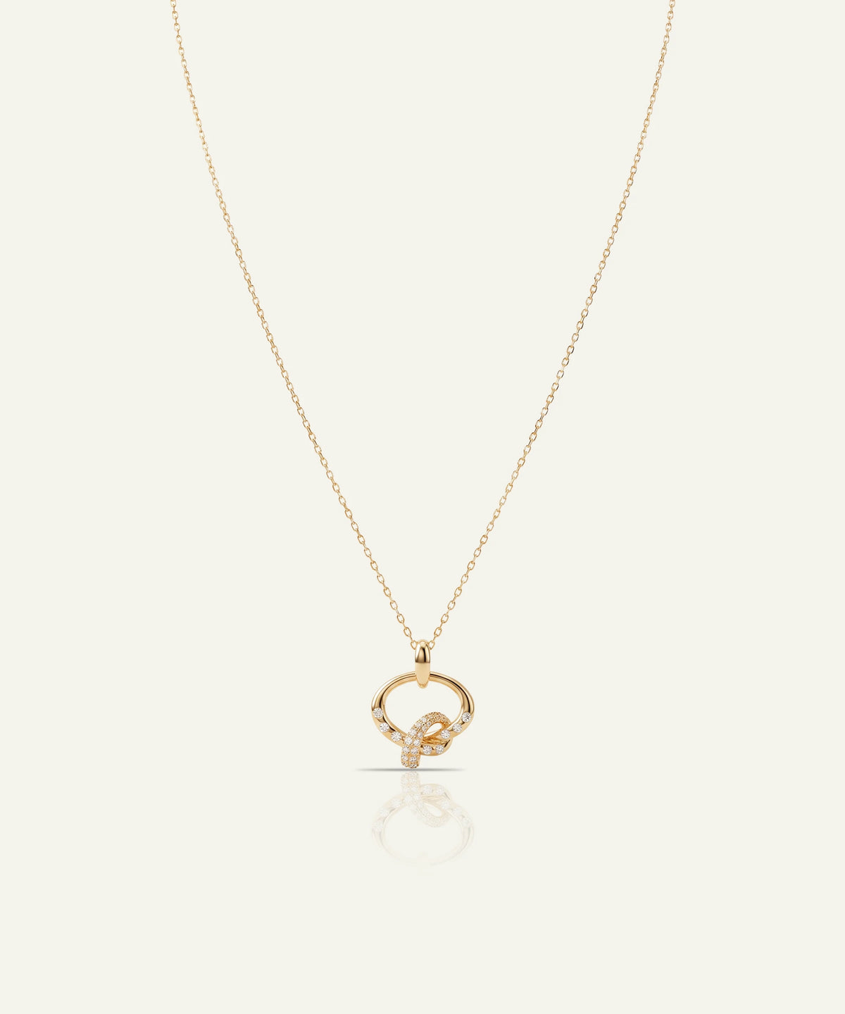 A delicate gold chain necklace featuring a circular pendant with a central knot design, accented by small clear stones, displayed on a light cream background with a soft reflection below.
