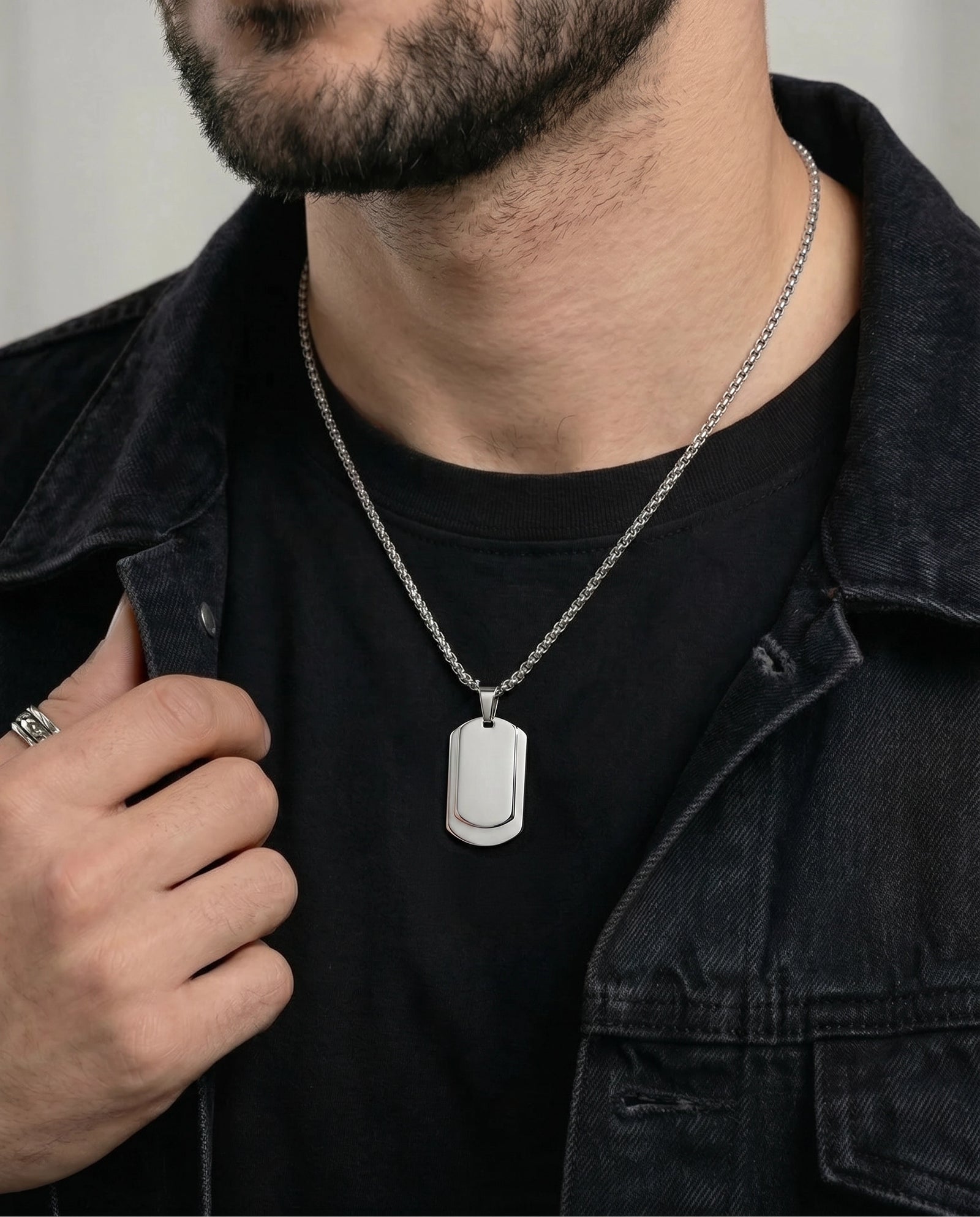 A close-up of a man wearing a silver dog tag pendant necklace over a black t-shirt and dark denim jacket.