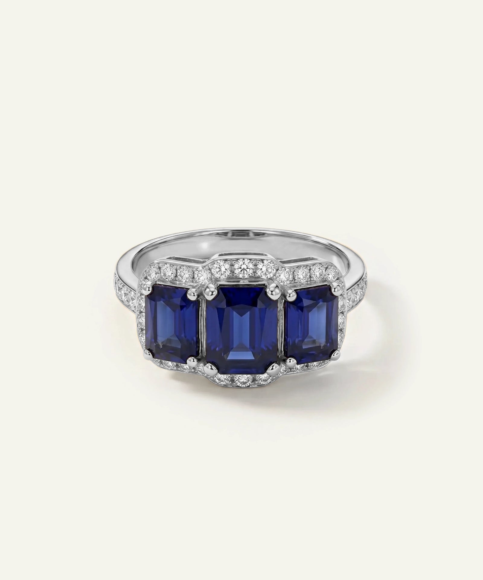 A silver-tone ring featuring three large rectangular deep blue sapphire gemstones, surrounded by small pavé-set clear stones, on a light cream background.
