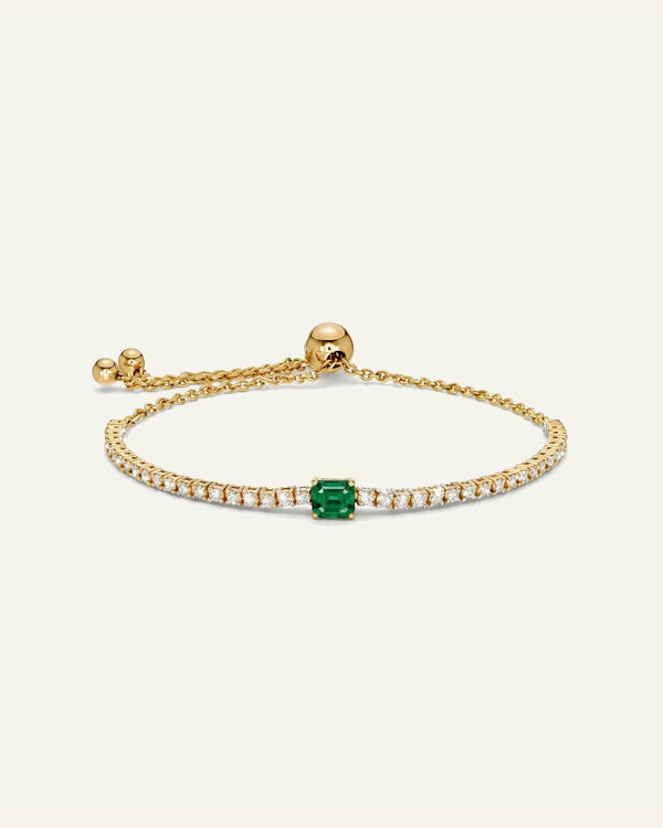 A delicate gold chain bracelet featuring a central emerald-cut green gemstone pavé-set with small clear stones, shown on a light cream background.