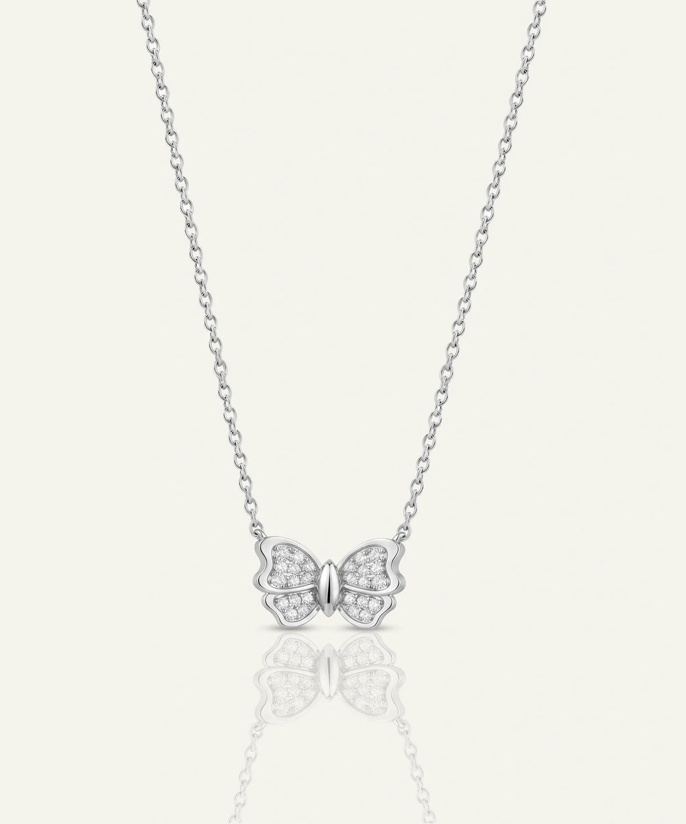A delicate silver chain necklace featuring a small butterfly pendant paved with tiny clear stones, centered on a light cream background with a soft reflection below.