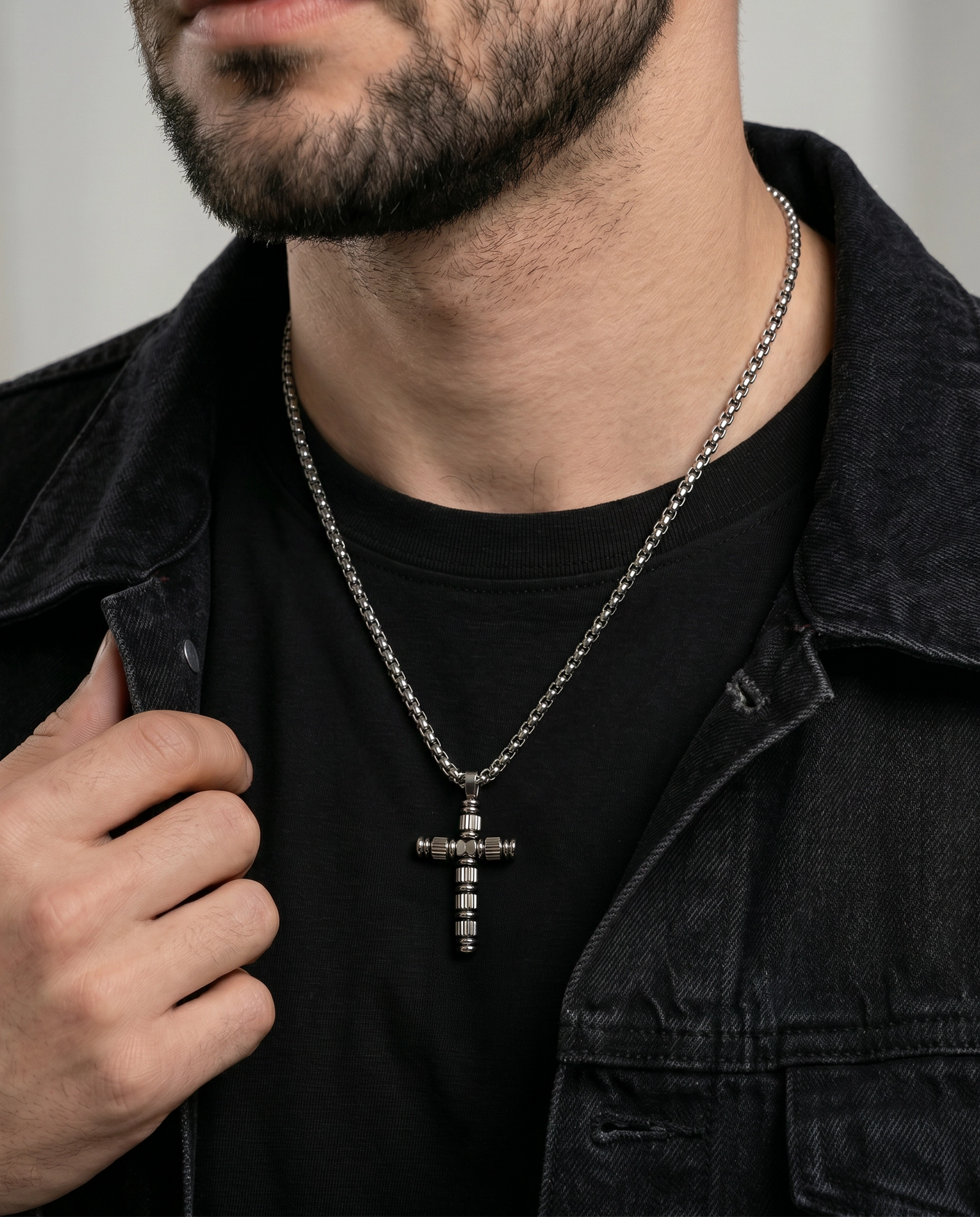 A close-up of a man wearing a silver-toned cross pendant necklace with a textured center, styled over a black t-shirt and a dark denim jacket.
