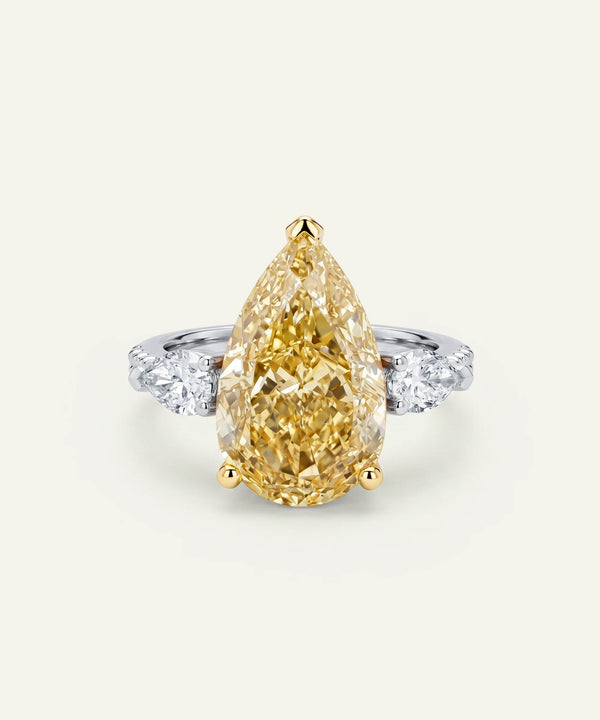 Pear-shaped fancy yellow diamond engagement ring set in yellow gold prongs, accented by round brilliant-cut white side diamonds on a white metal band. The ring is displayed from the front against a light neutral background.