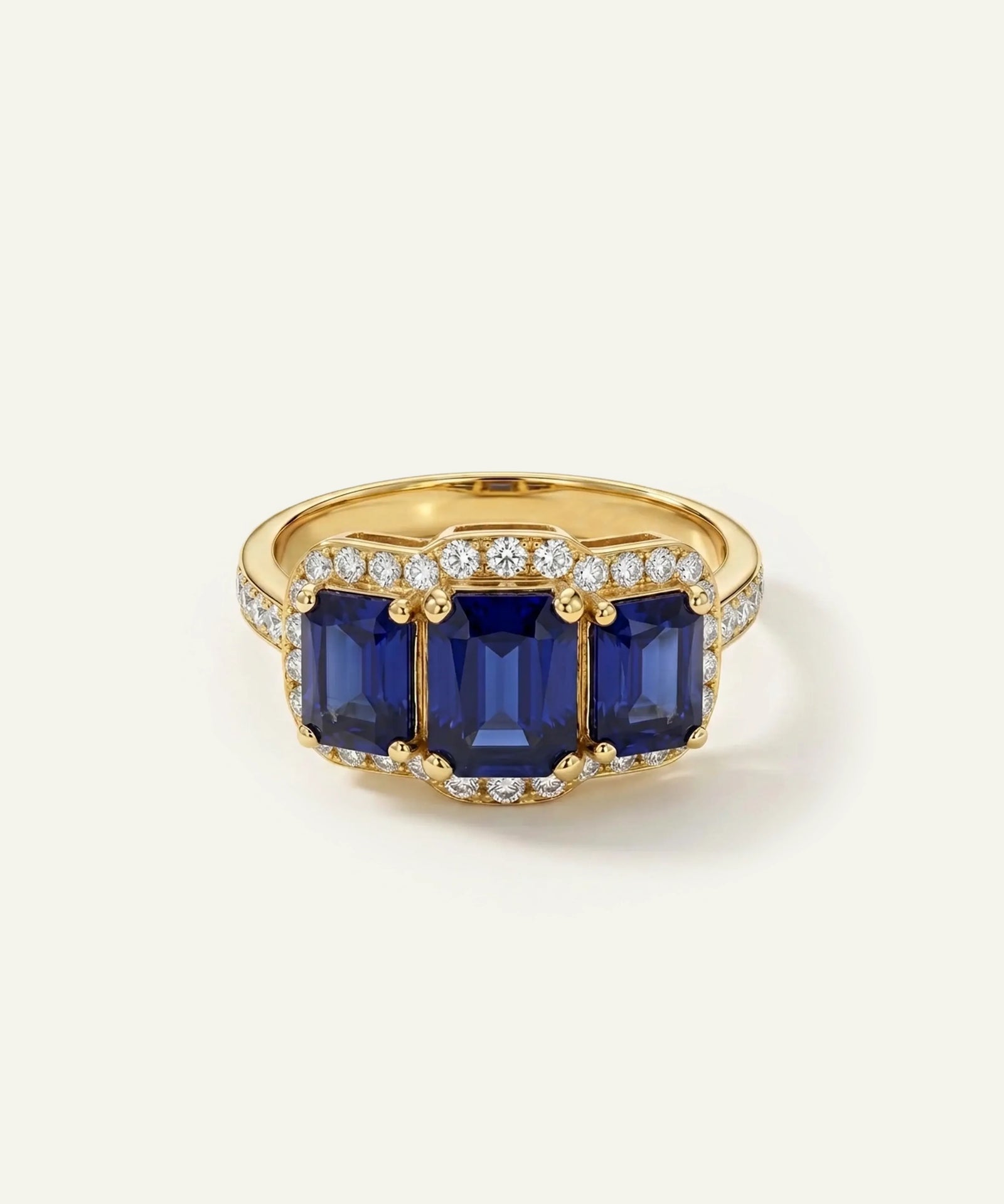 A yellow gold ring featuring three large rectangular blue sapphire gemstones, surrounded by small pavé-set clear stones, on a light cream background.