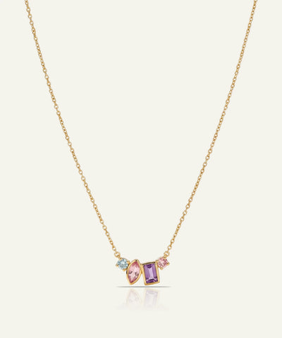 A delicate gold chain necklace featuring a small cluster pendant with a marquise-cut pink gemstone, a rectangular purple gemstone, and a tiny light blue gemstone, set against a light cream background.
