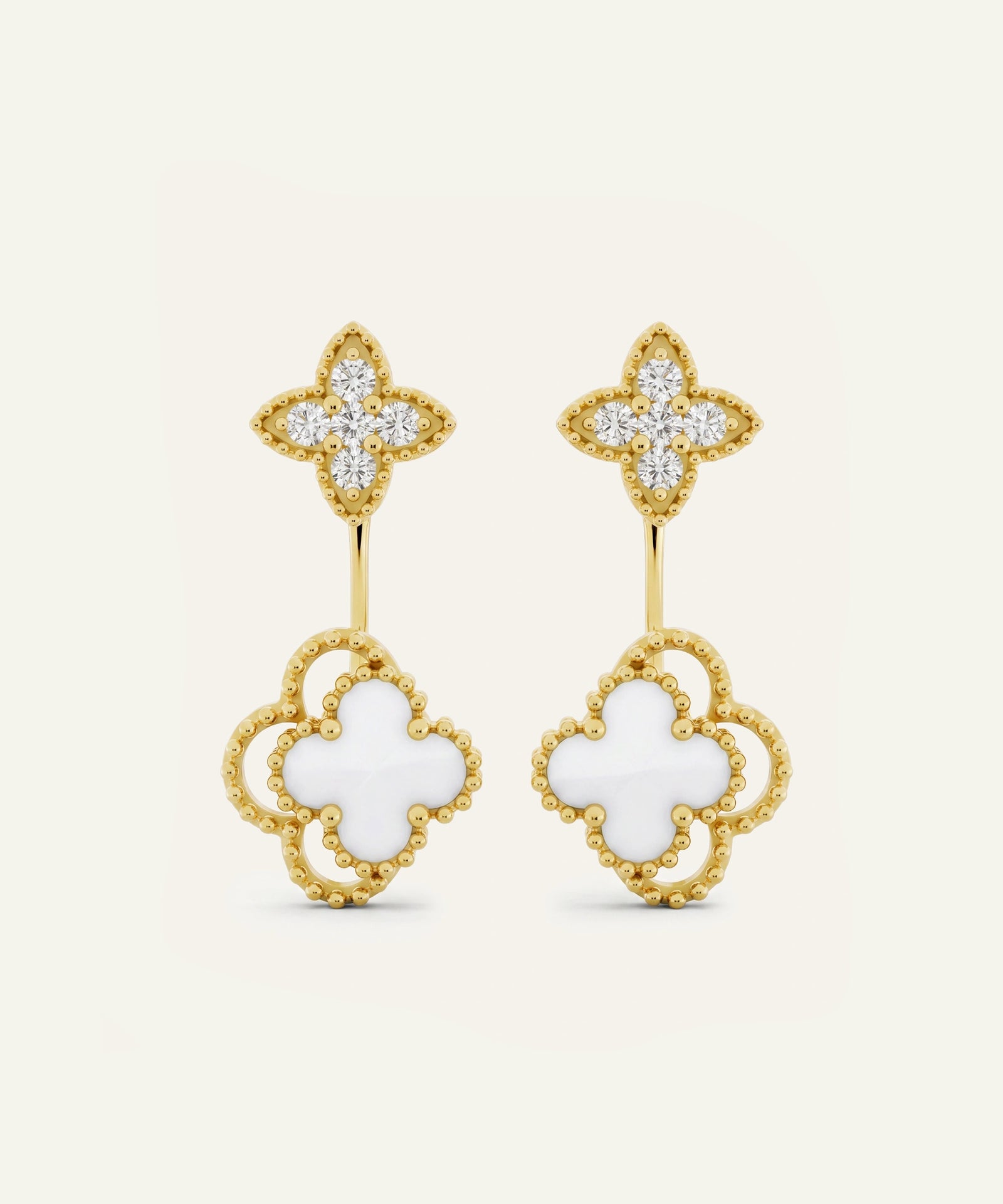A pair of gold and white clover drop earrings with pavé-set clear stones on the top clovers and openwork gold clover shapes with white inlay on the bottom, shown on a light cream background.