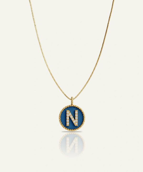 A gold chain necklace with an oval pendant featuring a blue enamel background and a raised gold letter 'N' outlined with small clear stones, shown on a light cream background with a soft reflection below.