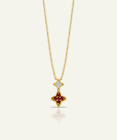 A gold chain necklace with a double-pendant design, featuring a small diamond-encrusted cross at the top and a larger gold cross pendant set with deep red gemstones below, shown on a light cream background.