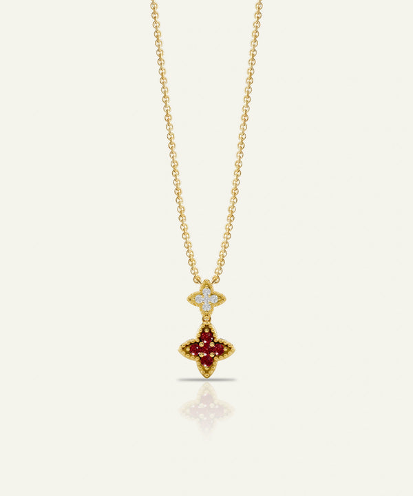 A gold chain necklace with a double-pendant design, featuring a small diamond-encrusted cross at the top and a larger gold cross pendant set with deep red gemstones below, shown on a light cream background.