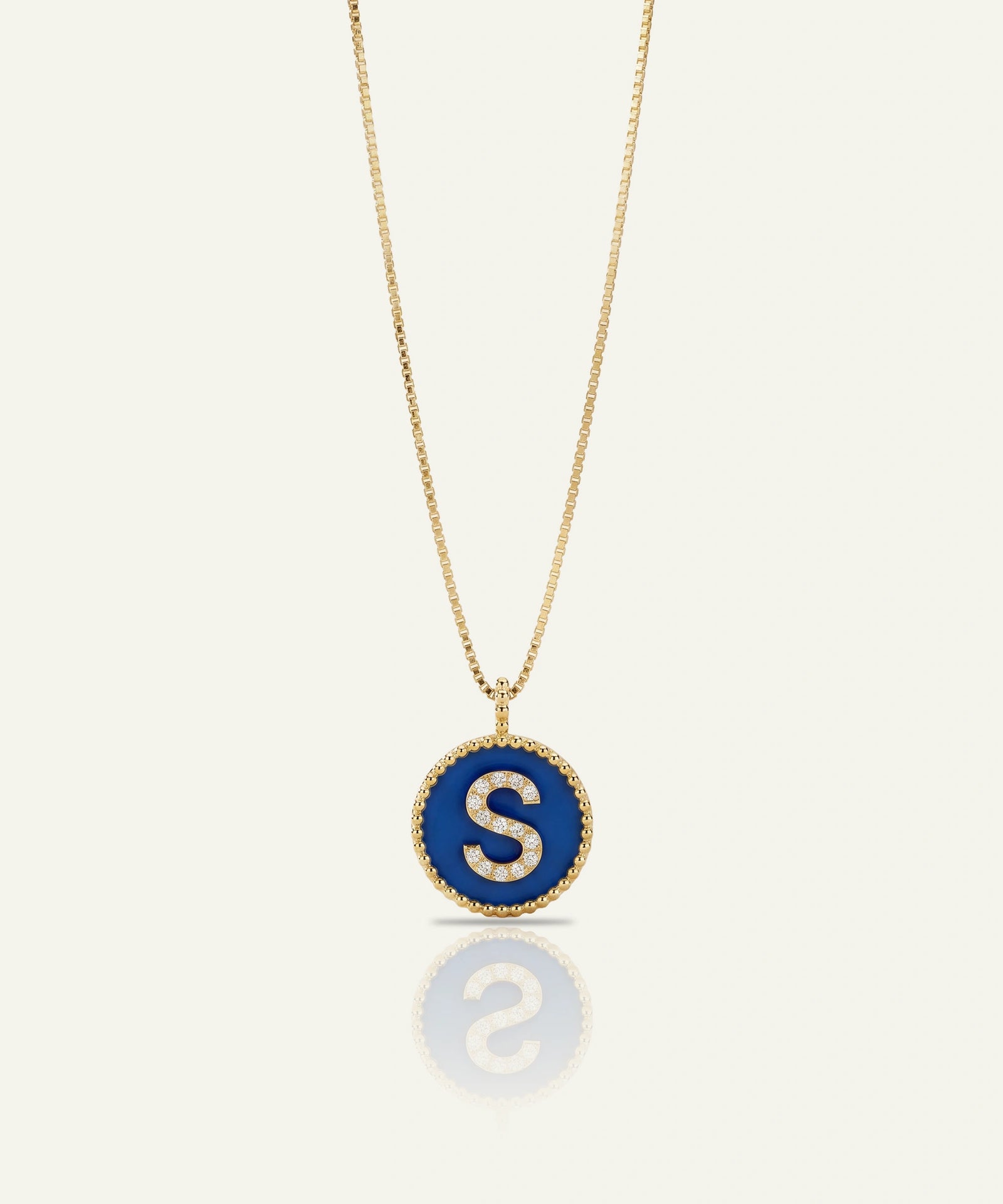 A gold chain necklace with a circular pendant featuring a deep blue enamel background and a white capital letter 'S' in the center, bordered by small clear stones, shown on a light cream background with a soft reflection below.