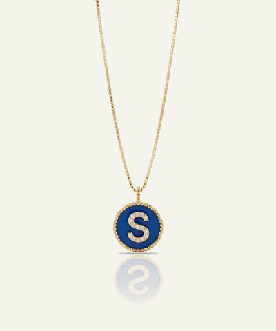 A gold chain necklace with a circular pendant featuring a deep blue enamel background and a white capital letter 'S' in the center, bordered by small clear stones, shown on a light cream background with a soft reflection below.