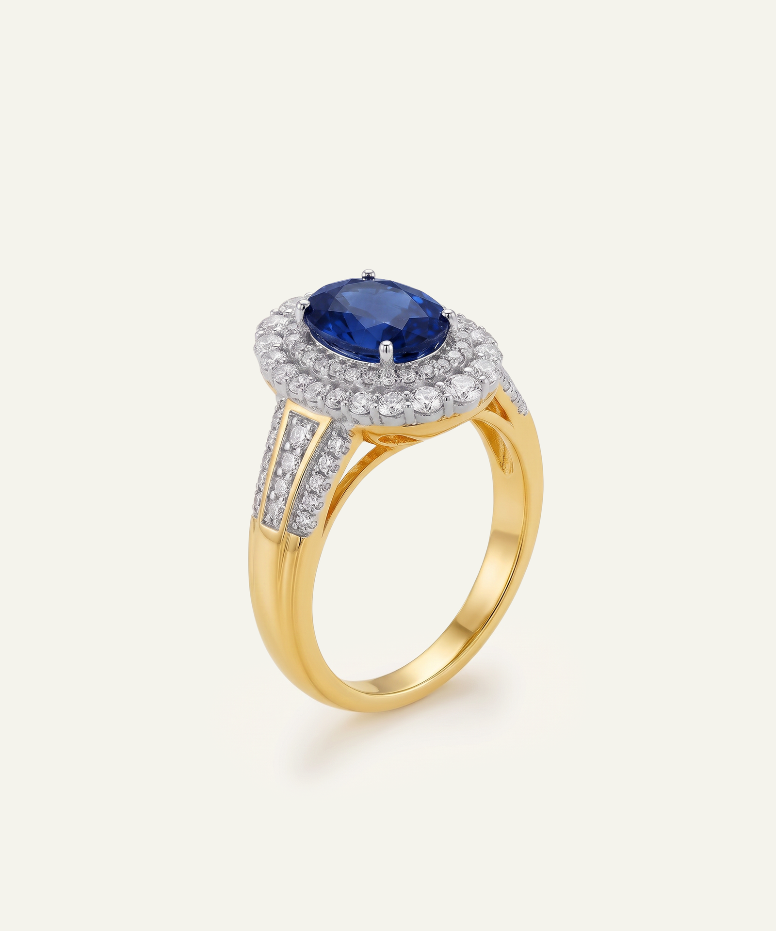 A yellow gold ring featuring a round-cut deep blue sapphire center stone surrounded by a halo of small clear stones, with additional clear stones set along the shoulders, shown on a light cream background.
