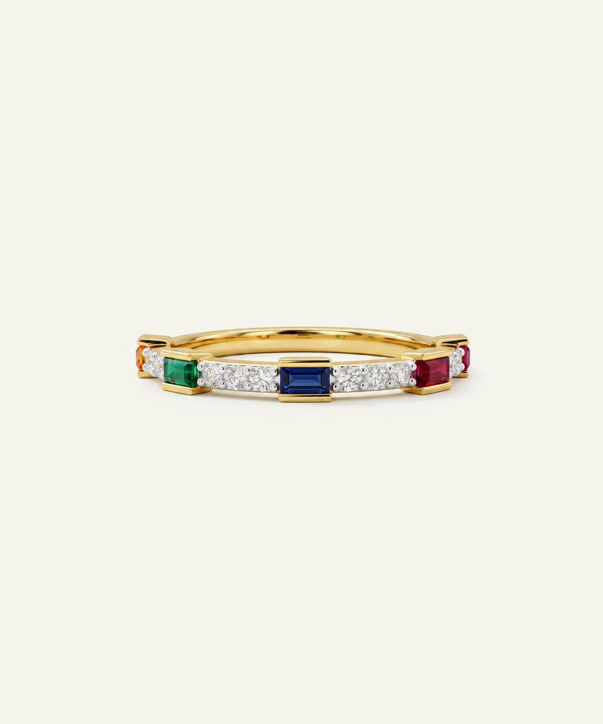 A gold band ring featuring a row of colorful gemstones, including emerald-cut blue, rectangular green, and round red stones, accented by rows of small clear stones, set against a light cream background.