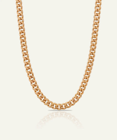 A gold-toned curb chain necklace displayed in a U-shape against a light cream background with a soft reflection below.