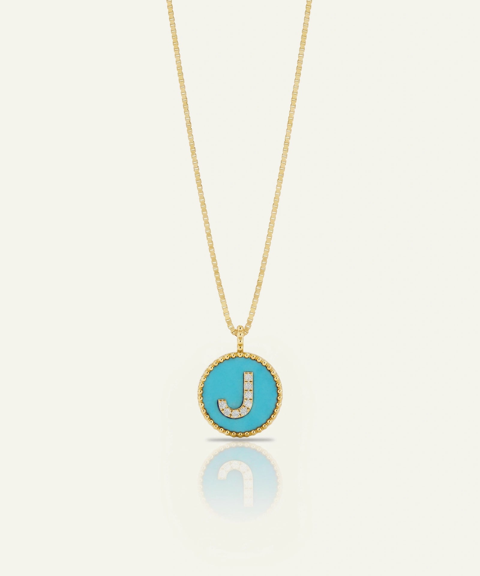 A gold chain necklace with a round turquoise pendant featuring a gold border and a letter 'J' paved with small clear stones, shown on a light cream background with a soft reflection below.