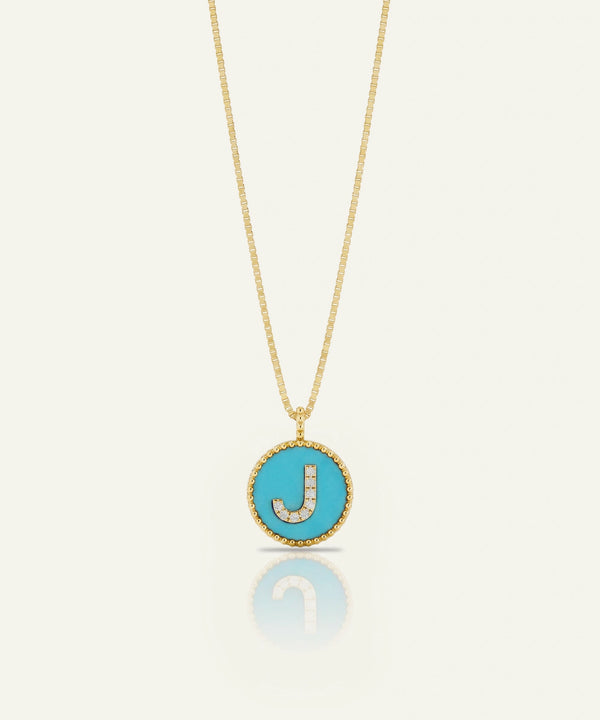 A gold chain necklace with a round turquoise pendant featuring a gold border and a letter 'J' paved with small clear stones, shown on a light cream background with a soft reflection below.