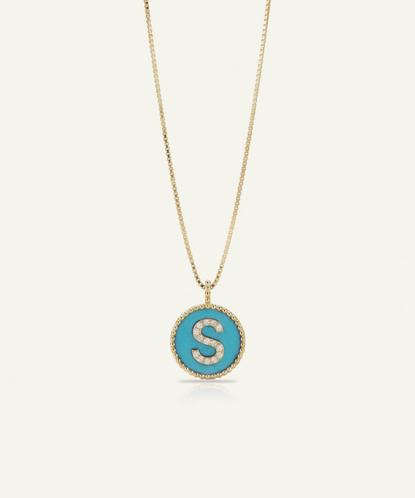 A gold chain necklace with a round pendant featuring a turquoise blue background and a gold letter 'S' outlined with small clear stones, shown on a light cream background.