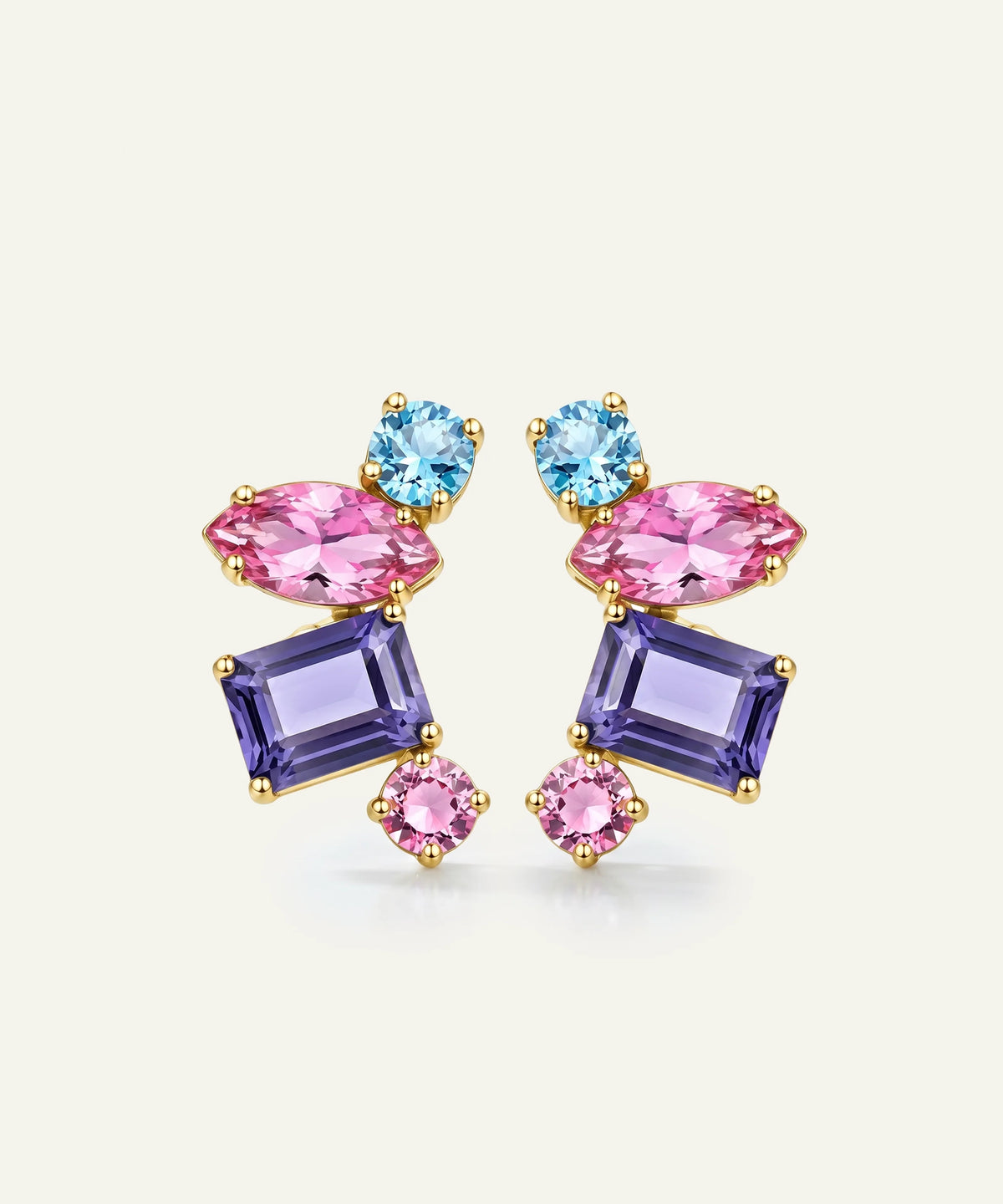 A pair of gold-plated statement earrings featuring marquise-cut pink crystals, rectangular purple crystals, and round light blue crystals, set against a light cream background.